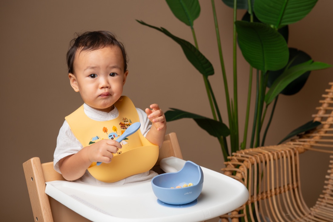 Toddler in a yellow Little Rei silicone bib eating from a blue suction bowl in a high chair