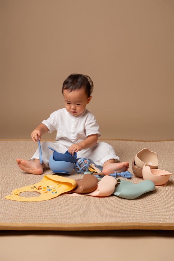 Baby sitting on a playmat surrounded by Little Rei silicone bowls, bibs and a spoon