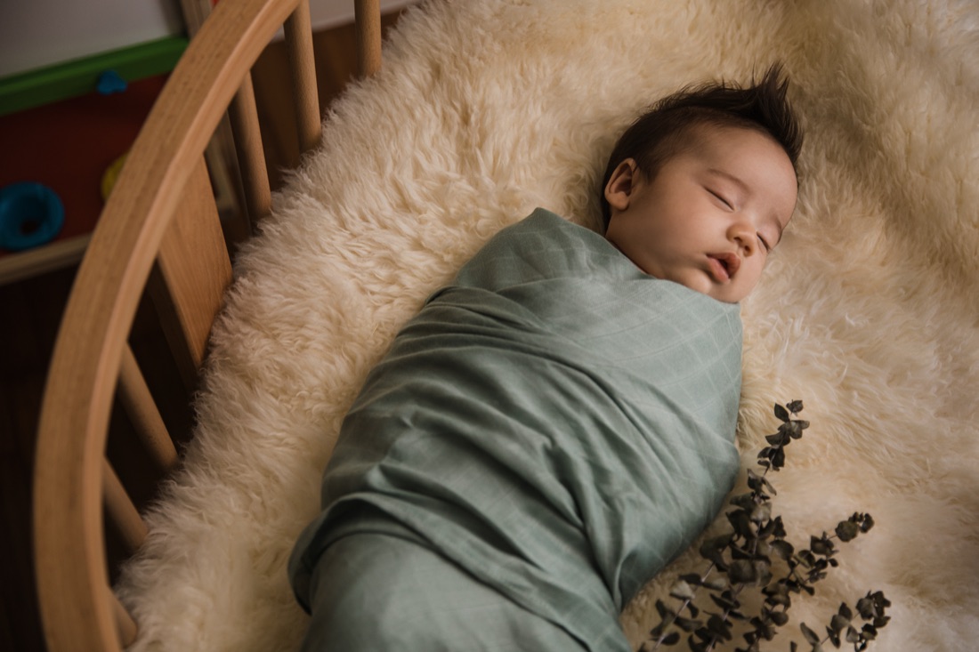 Newborn asleep in a cot, swaddled in a sage green Little Rei bamboo muslin